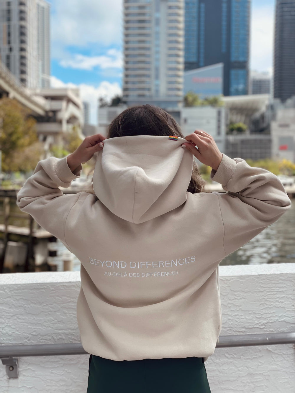 Personne de dos en ville portant le Heavyweight Hoodie Beige, broderie BEYOND DIFFERENCES au dos