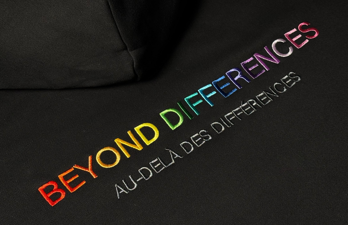 Gros plan de la broderie arc-en-ciel BEYOND DIFFERENCES / AU-DELÀ DES DIFFÉRENCES sur un hoodie noir