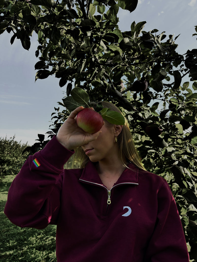 Person wearing the burgundy quarter-zip in an orchard, holding an apple